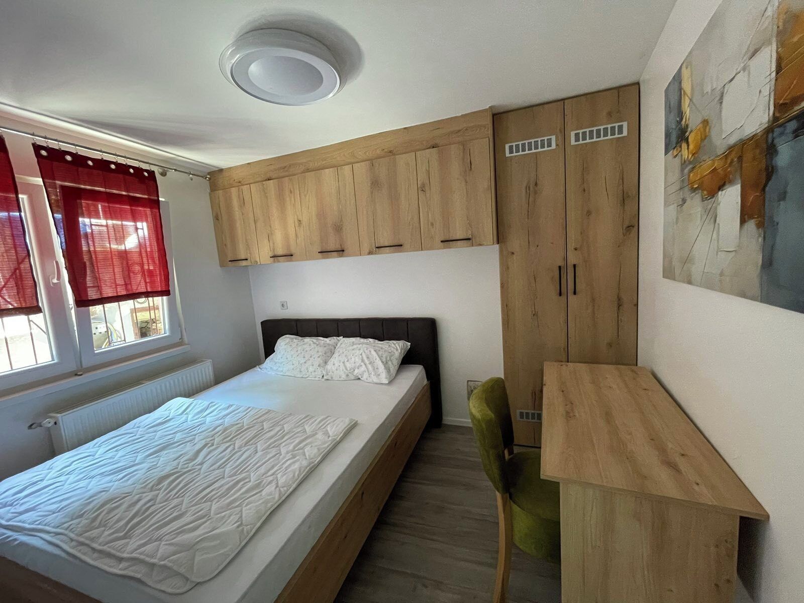 A bedroom with a bed, a desk, and a chair. There is a window with red curtains and a radiator below it.
