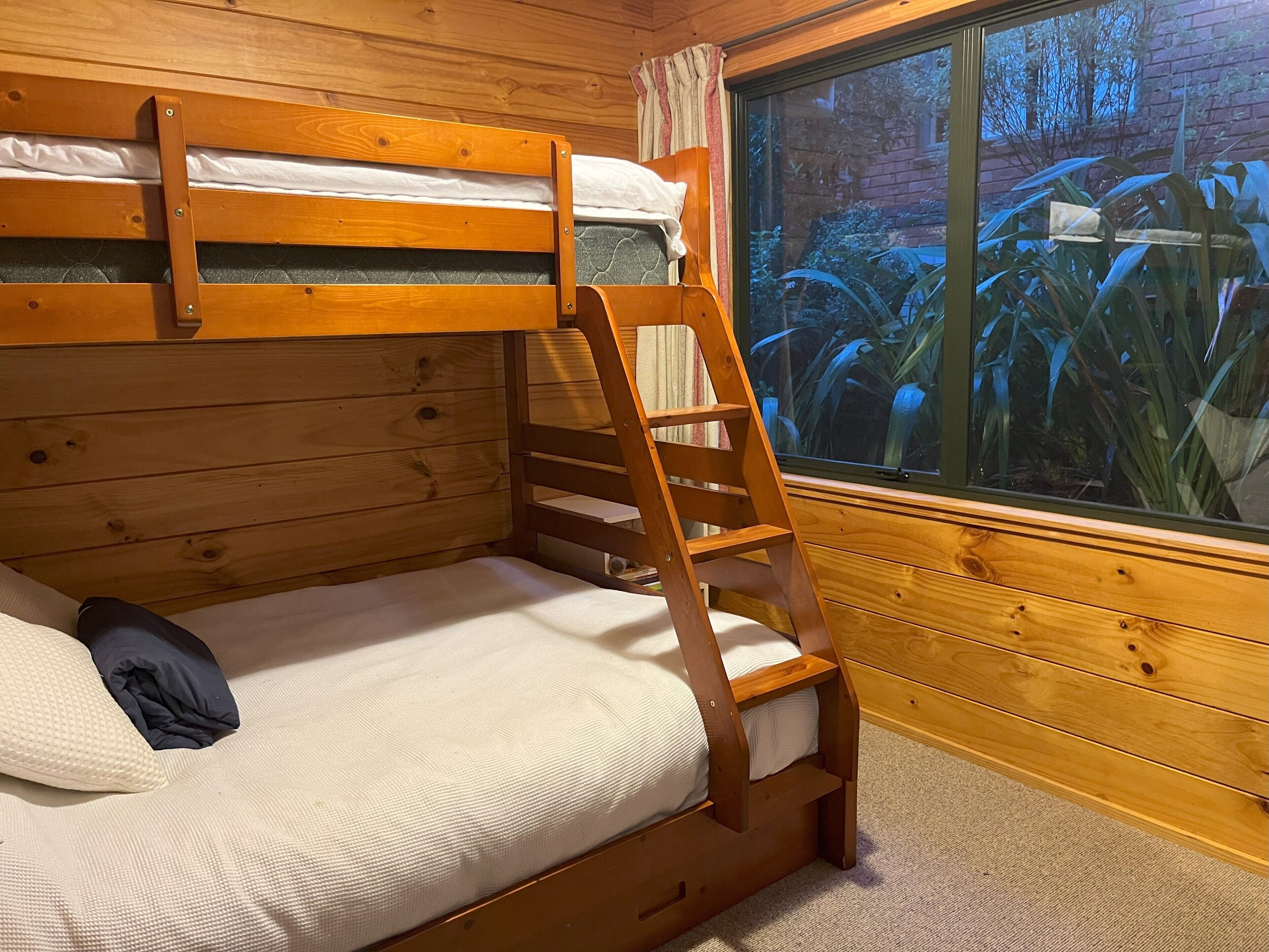 A bunk bed with a wooden ladder, a window with a view of plants, and a wooden wall.