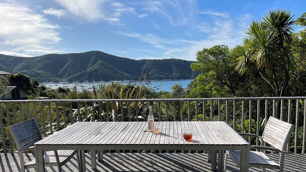 Una terraza de madera con una mesa y sillas, con vista a una bahía con barcos y colinas.
