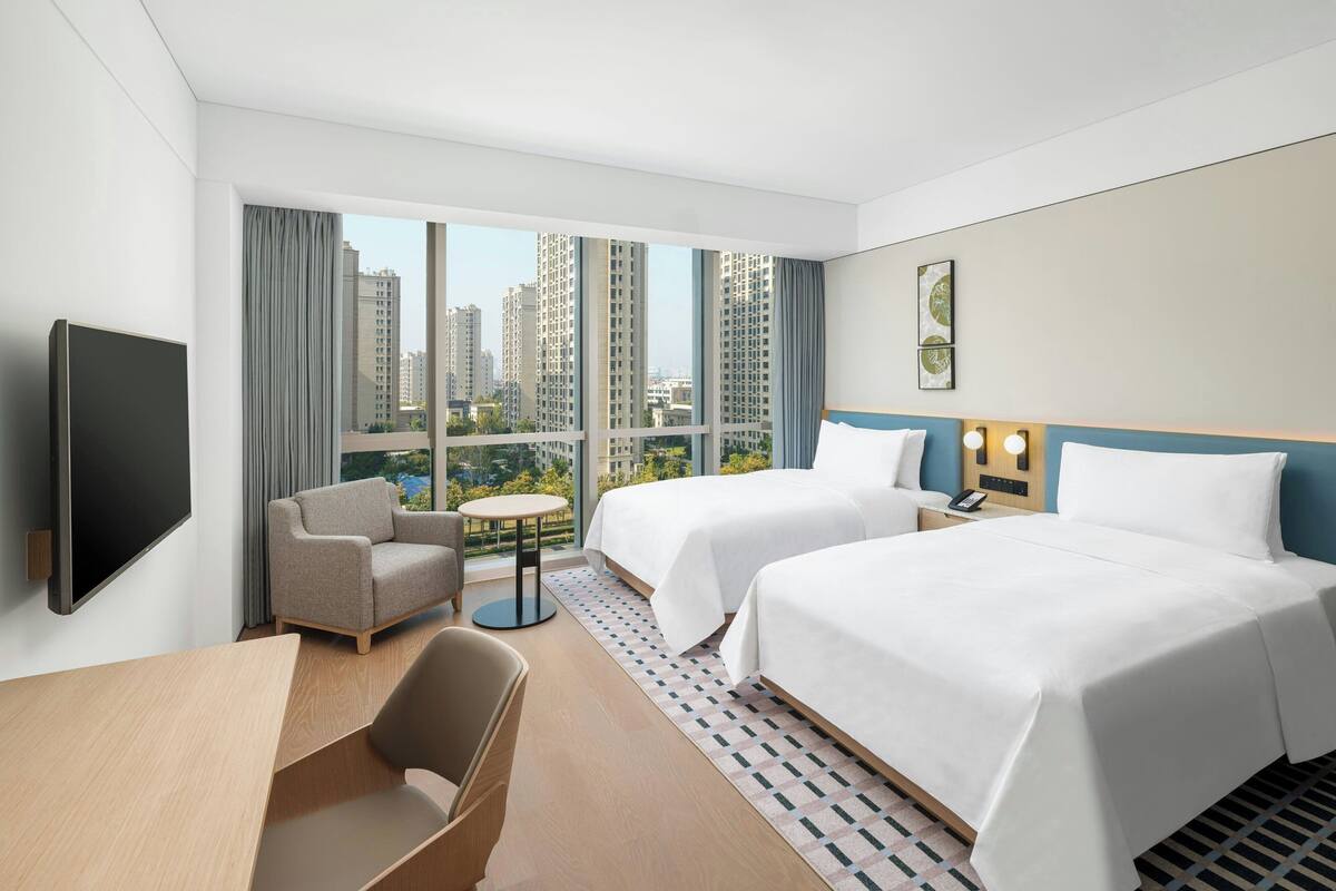 Habitación de hotel con dos camas, televisión, escritorio y vistas a edificios altos.