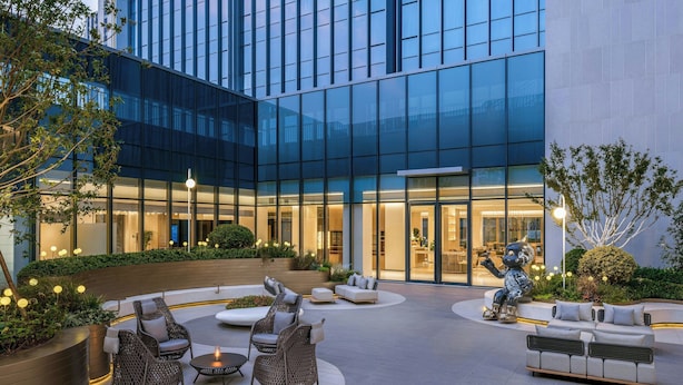 Une cour d’hôtel moderne avec des espaces de détente en plein air, une sculpture et une façade en verre.