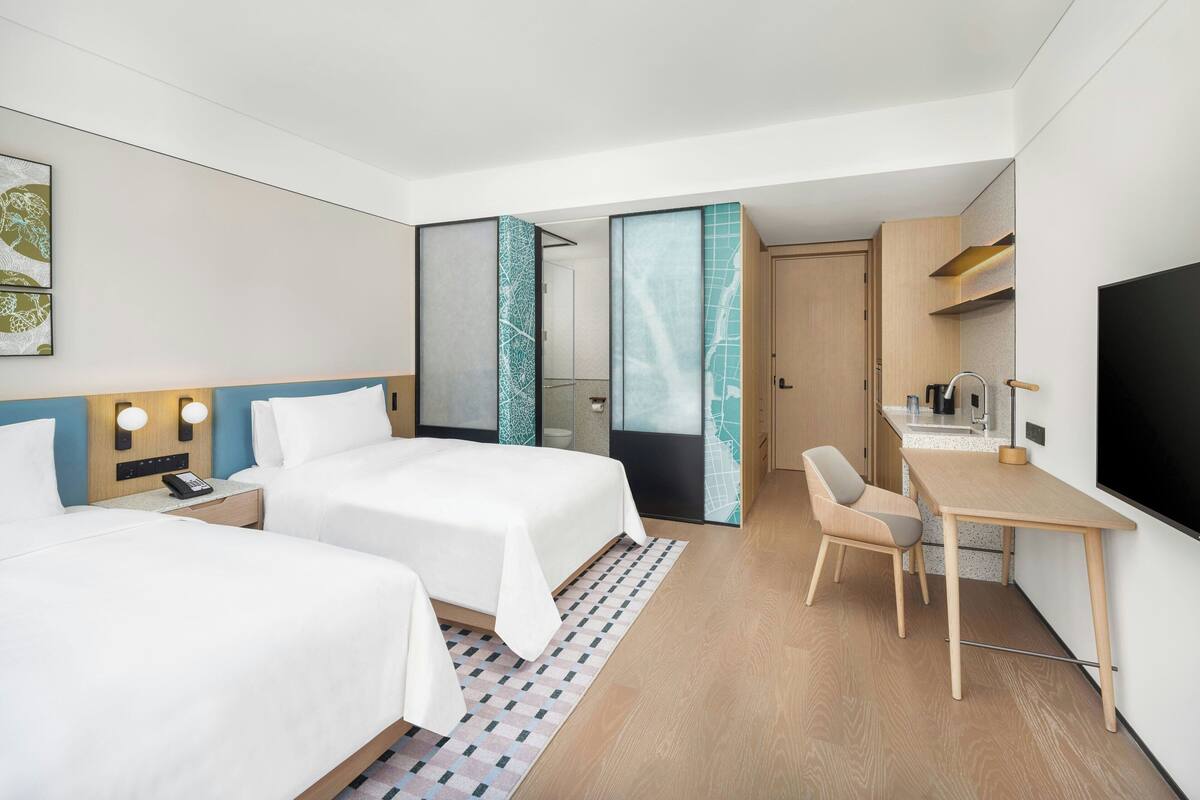 Habitación de hotel con dos camas, un escritorio, una silla, un televisor y un baño con puerta de vidrio.