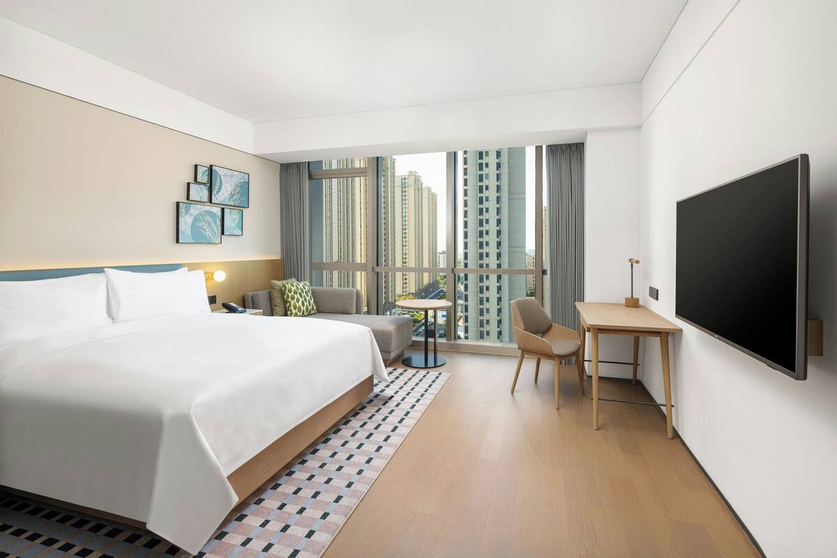 Habitación de hotel con una cama grande, un escritorio, una silla, un televisor y vistas de edificios altos a través de la ventana.