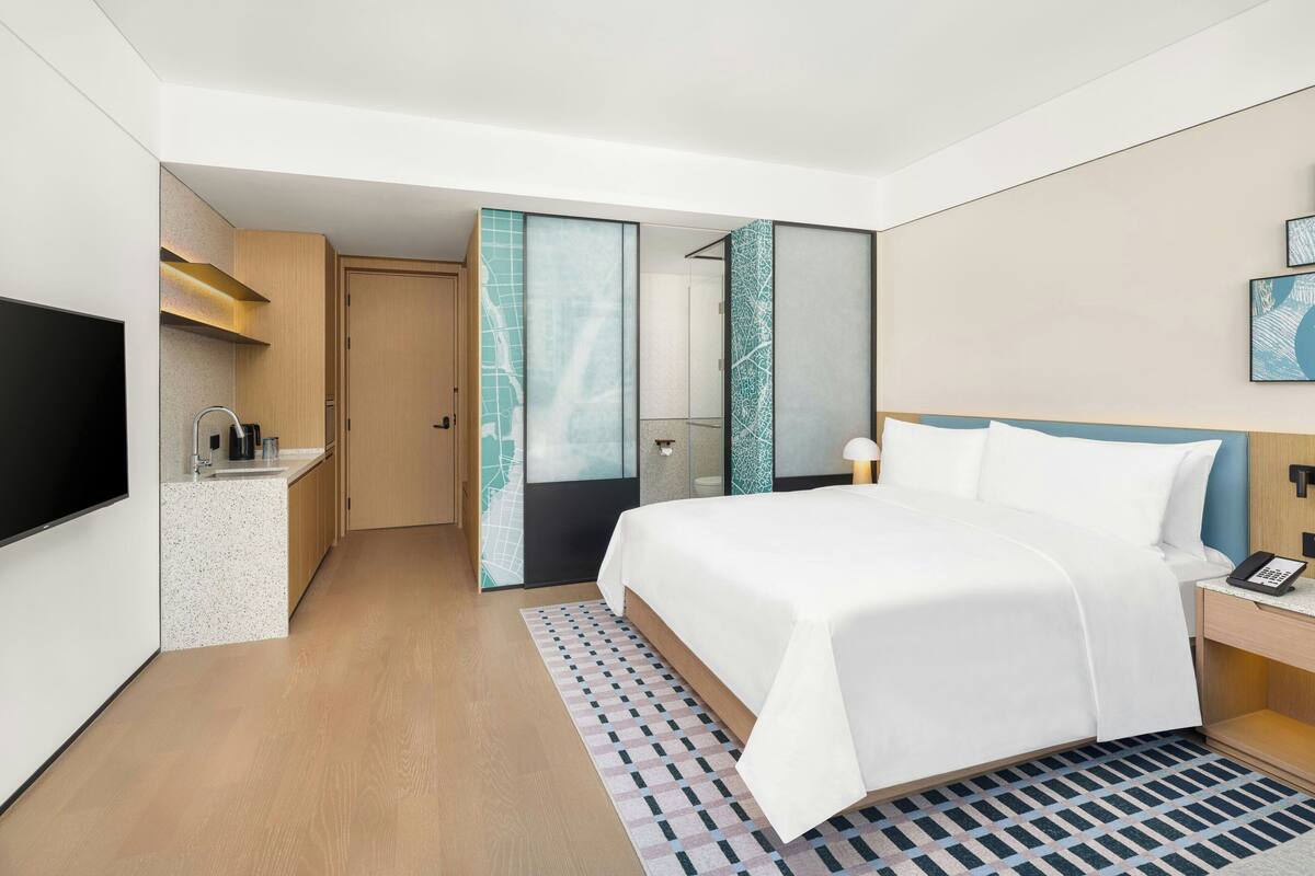 Una habitación de hotel moderna con una cama grande, una mesita de noche con teléfono, un televisor de pantalla plana, una pequeña cocina y un baño con puerta de vidrio.
