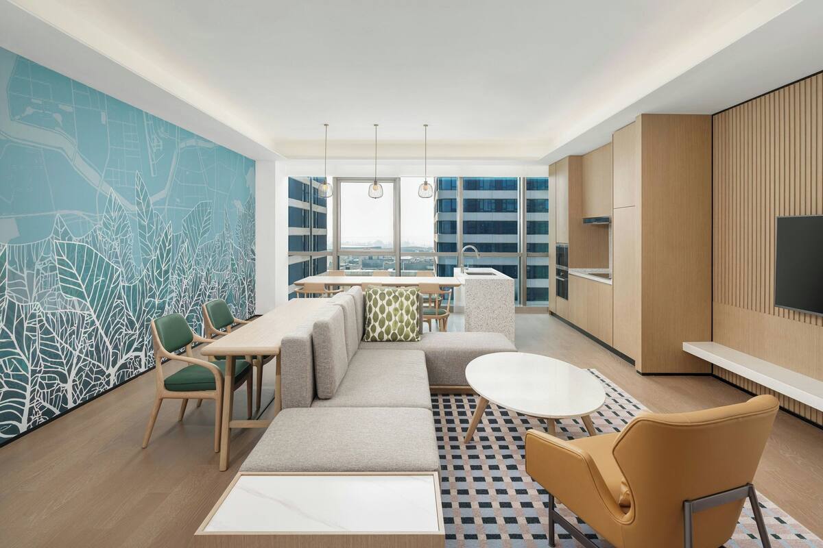 Habitación de hotel moderna con zona de comedor, sofá, una mesa redonda y un ventanal con vistas a la ciudad.