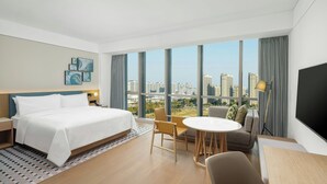 Une chambre d’hôtel moderne avec un grand lit, une table à manger et un canapé. De grandes fenêtres offrent une vue sur la ville.