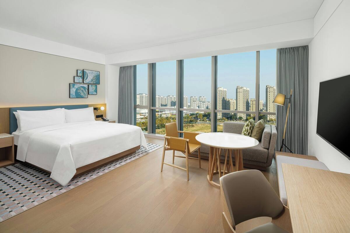 Una habitación de hotel moderna con una cama grande, una mesa de comedor y un sofá. Amplias ventanas ofrecen vistas a la ciudad.