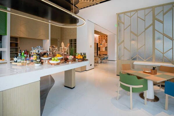 A modern interior with a long counter displaying various bottles and fruits, a seating area with a table and chairs, and a kitchen in the background.