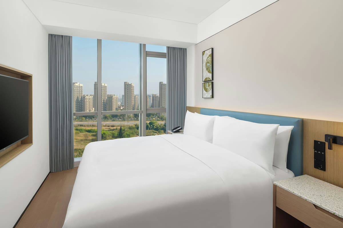 Habitación de hotel con una cama grande, televisión, mesita de noche y vistas a la ciudad a través de amplios ventanales.