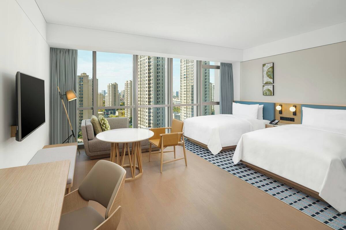 Habitación de hotel con dos camas, televisión, escritorio y vistas a edificios altos.