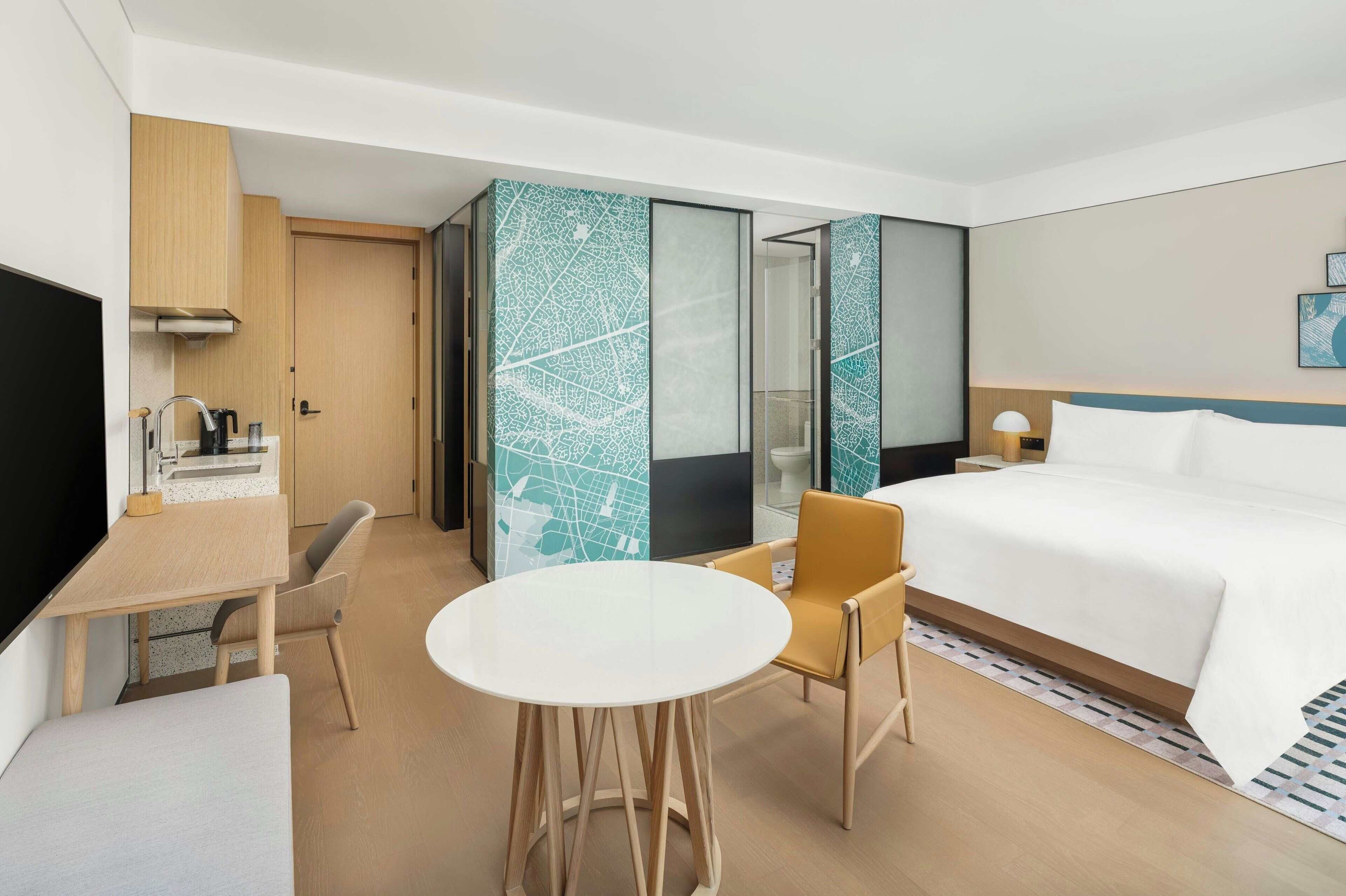 A hotel room with a bed, a desk, a chair, a round table, and a bathroom with a glass door.