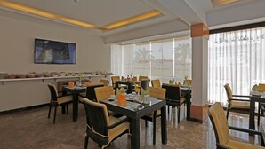 A hotel dining area with multiple tables and chairs, a television mounted on the wall, and large windows with a view of palm trees. - Hotelier Suites by Cantonal (Riyadh)