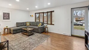 A modern living room with a sectional sofa, a coffee table, and a rug. There are two windows with blinds, a door, and a framed picture on the wall. - Packer Pilgrimage 1 - Sunroom + Pet Friendly (Green Bay)
