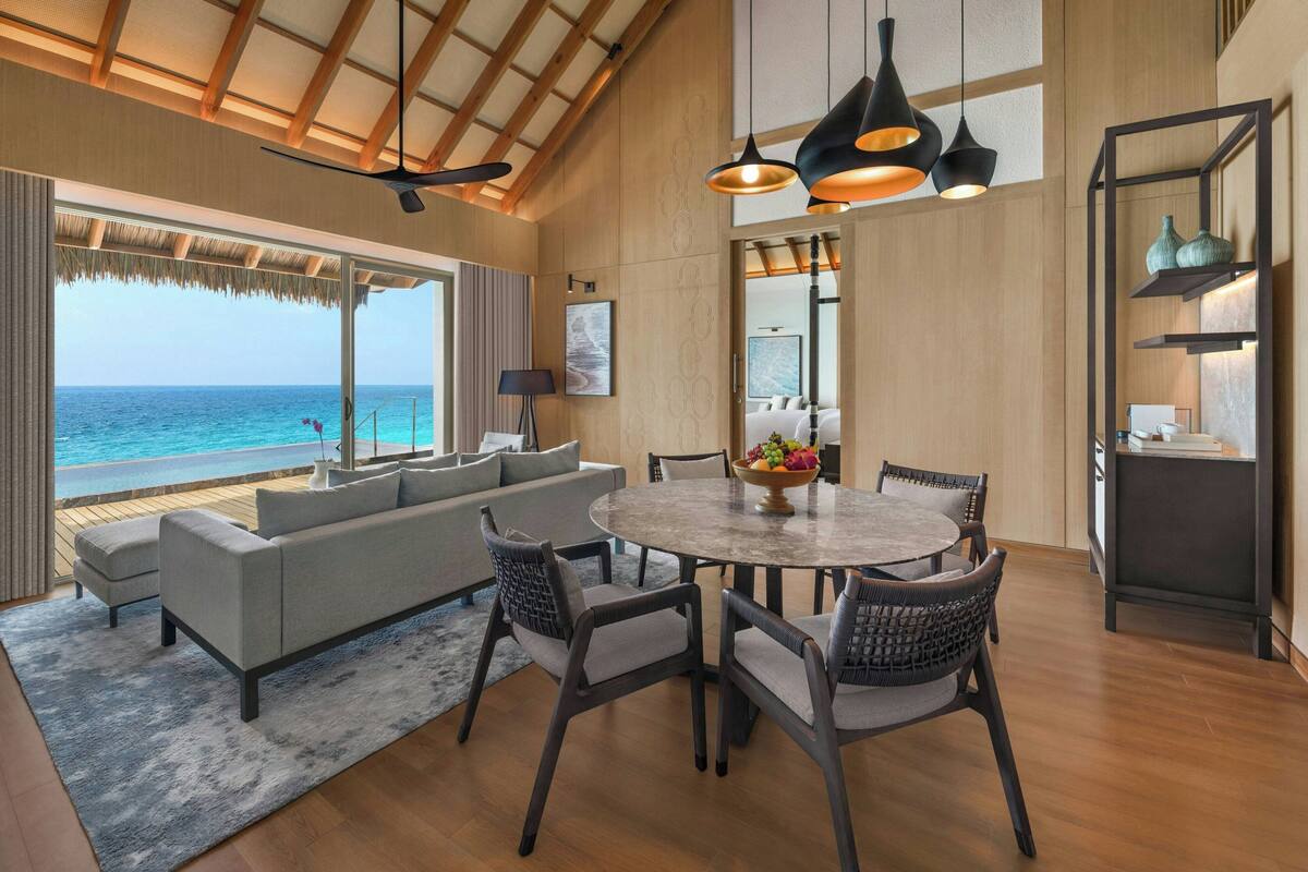 A spacious living area with a large window overlooking the ocean, a round dining table with chairs, and a modern chandelier.