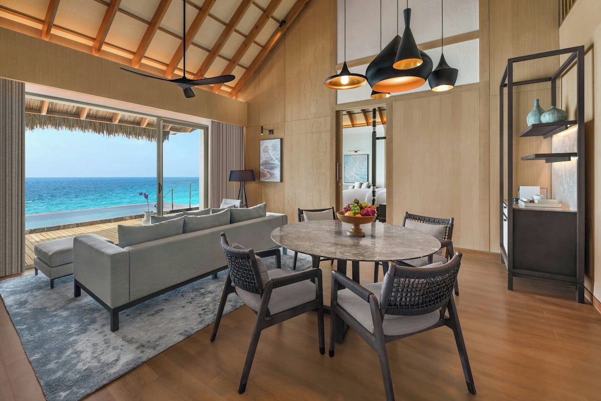A spacious living area with a large window overlooking the ocean, a round dining table with chairs, and a modern chandelier.
