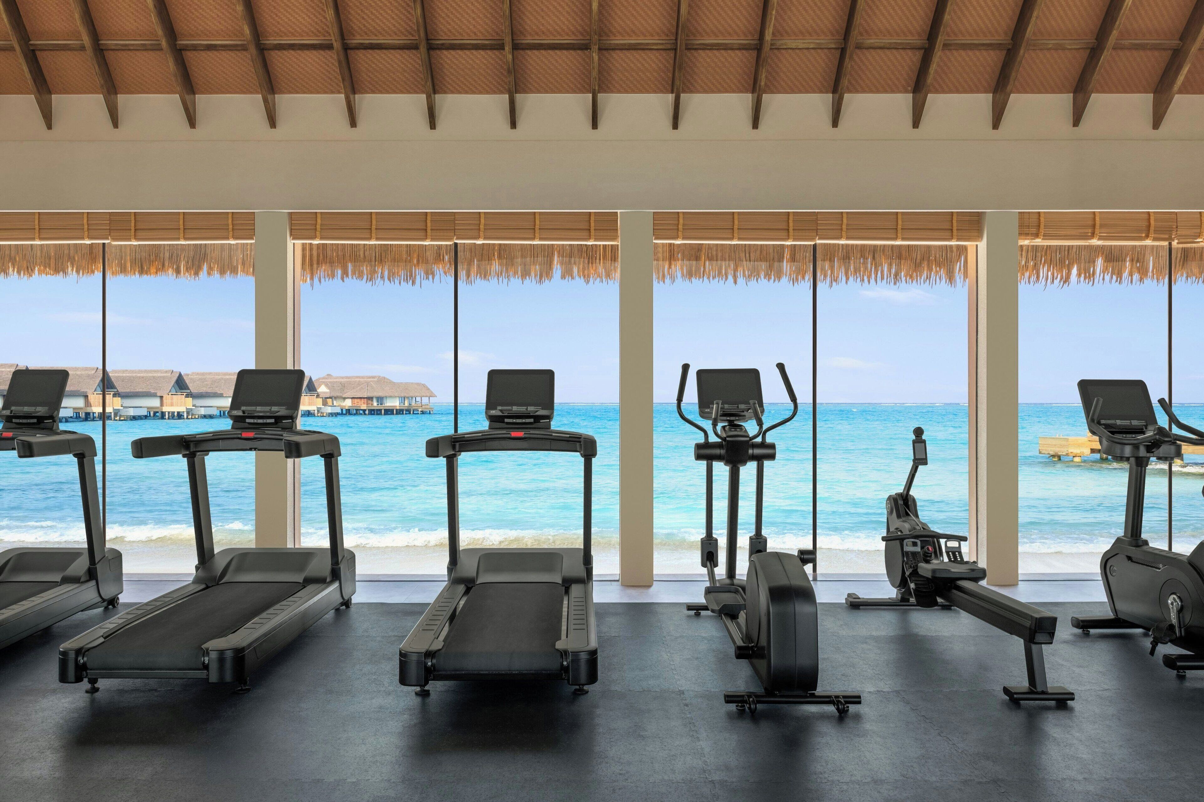 Une salle de sport équipée de tapis de course et d’appareils d’exercice, avec vue sur une plage et des bungalows sur pilotis.