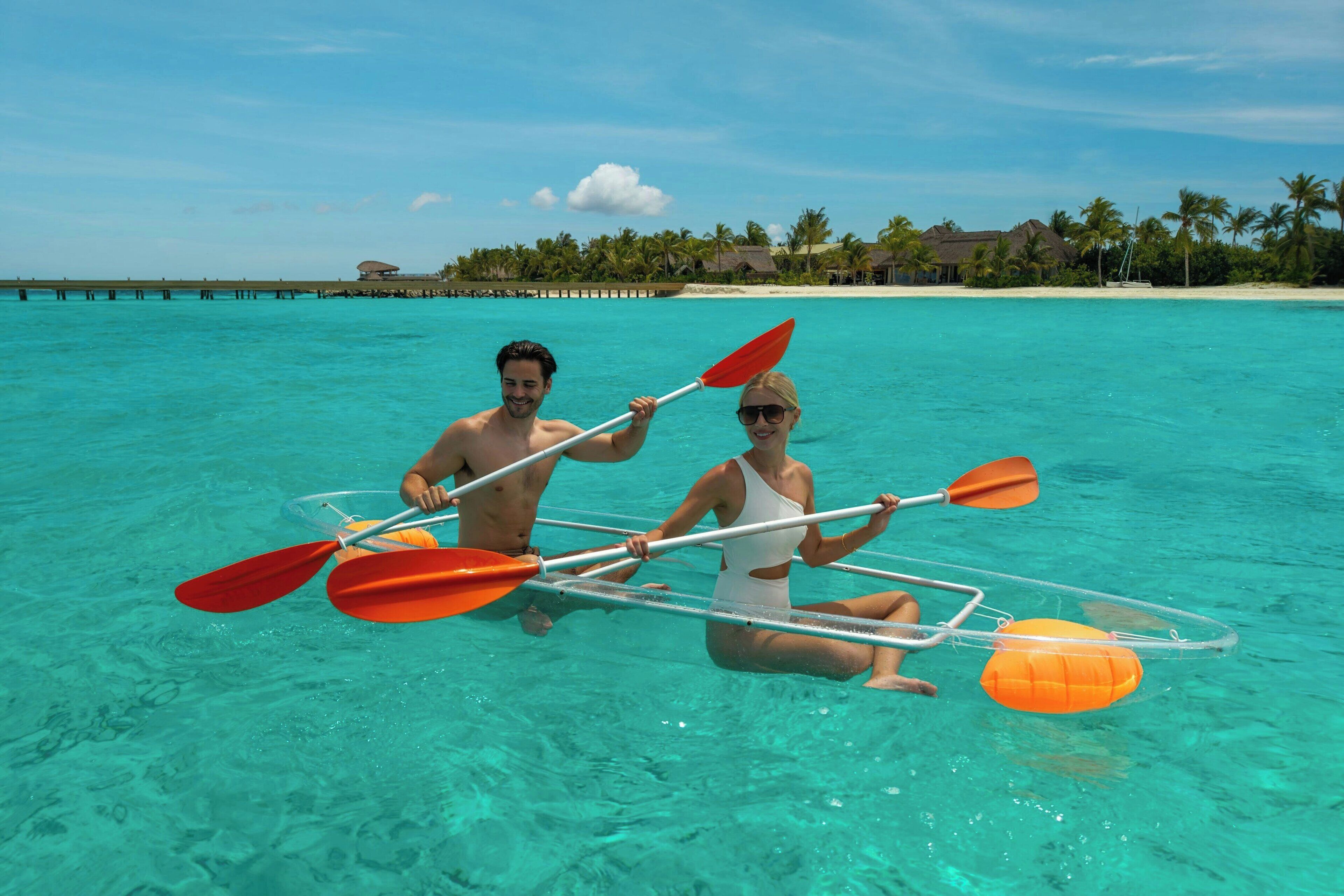 Two people kayaking in clear turquoise water with a beach and palm trees in the background.