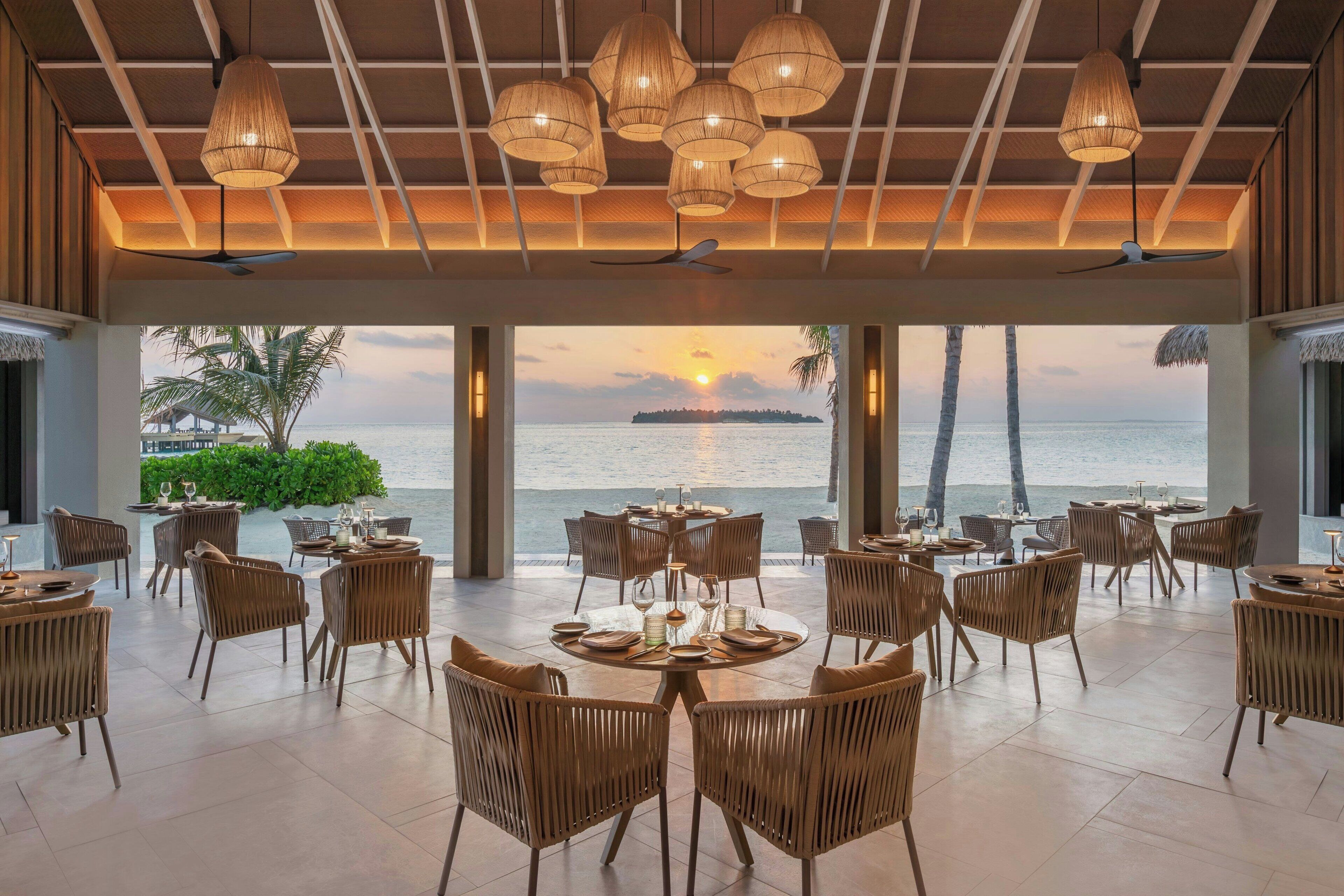 A spacious dining area with multiple tables and chairs, large windows offering a view of the sea and palm trees, and a ceiling with woven light fixtures.