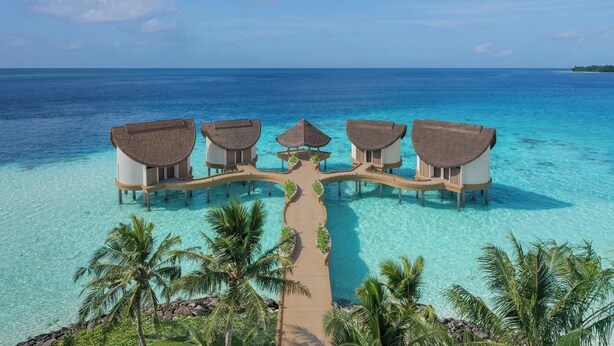 An aerial view of a resort with overwater bungalows connected by a wooden walkway, surrounded by clear turquoise waters and palm trees. - JW Marriott Maldives Kaafu Atoll Island Resort (Hathaa Finolhu)