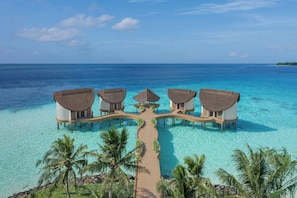 Vista aérea de un resort con bungalows sobre el agua, conectados por una pasarela de madera, rodeado de aguas turquesas cristalinas y palmeras.