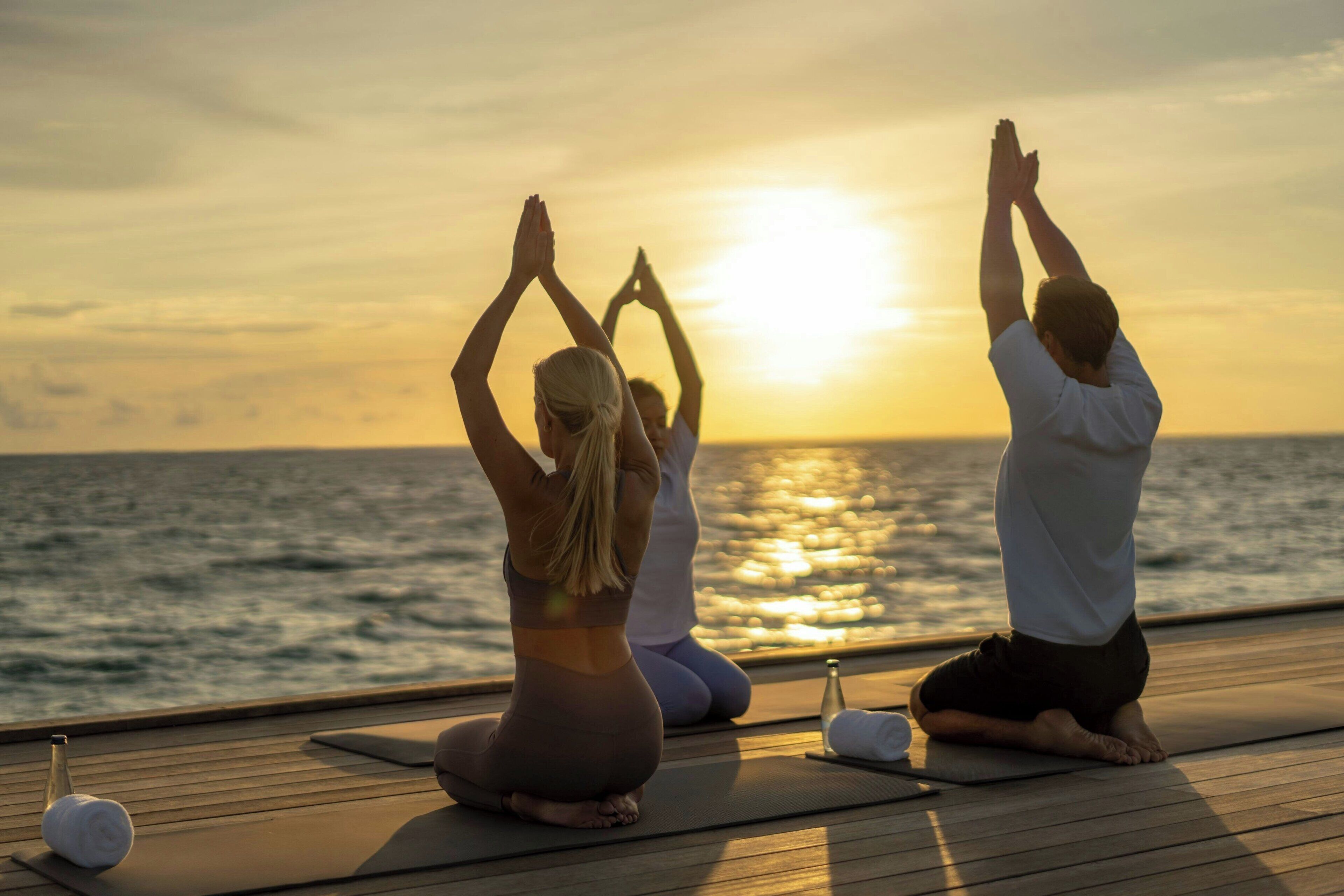 Trois personnes pratiquent le yoga sur une terrasse en bois, avec vue sur l’océan au coucher du soleil.