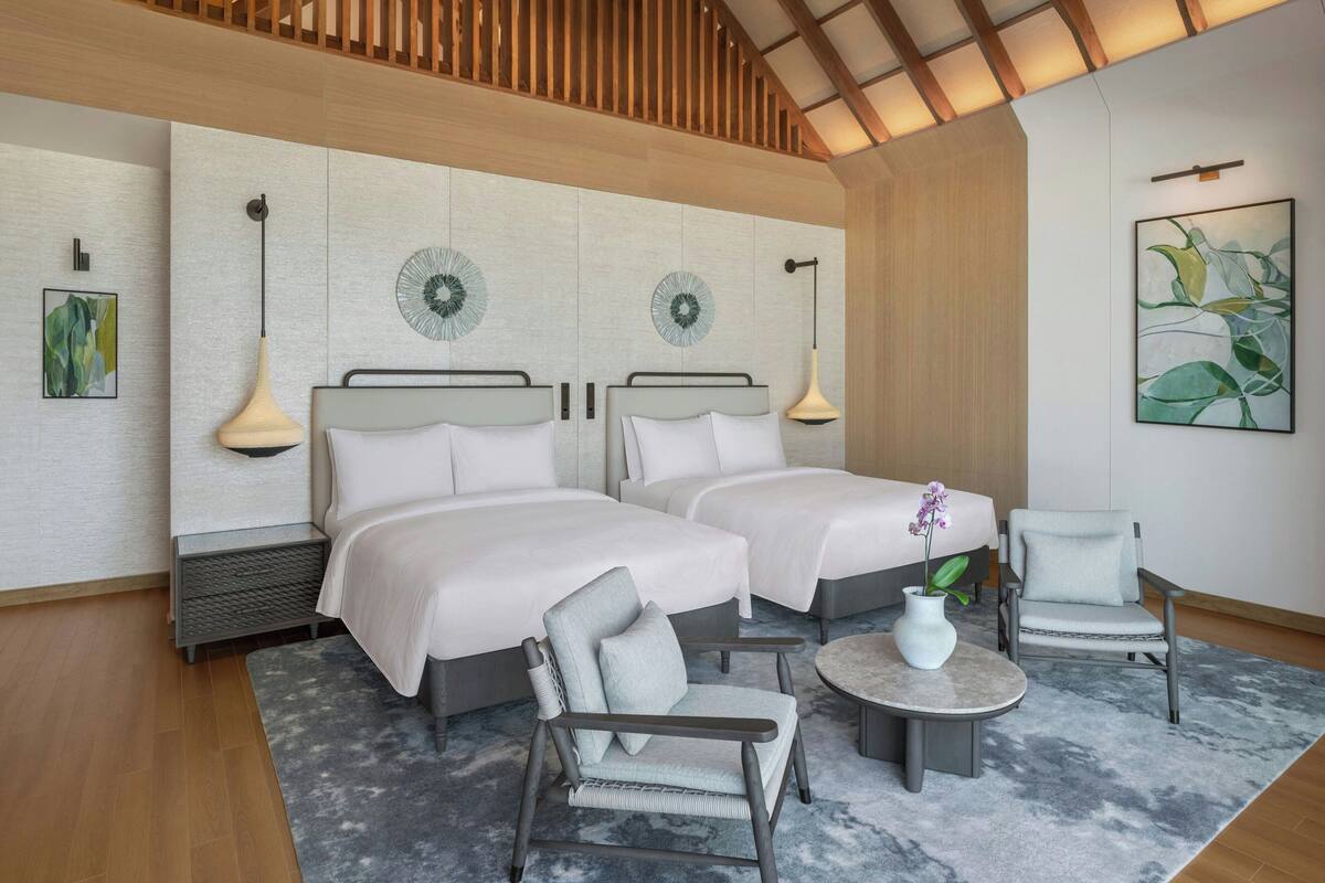 A hotel room with two beds, a chair, a small table, and a decorative plant.