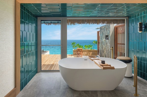 Un baño moderno con una bañera independiente, una terraza de madera con vista al océano y un entorno tropical con palmeras.