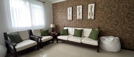 Sala de estar con un sofá blanco, un sillón de madera y una mesa de centro. Se observan tres obras de arte enmarcadas en la pared y una ventana con cortinas de encaje.