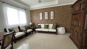 Ein Wohnzimmer mit einem weißen Sofa, zwei Sesseln, einem Holztisch und einem Esstisch mit Stühlen. Rechts befindet sich ein großer Schrank und eine Wand mit drei gerahmten Bildern.