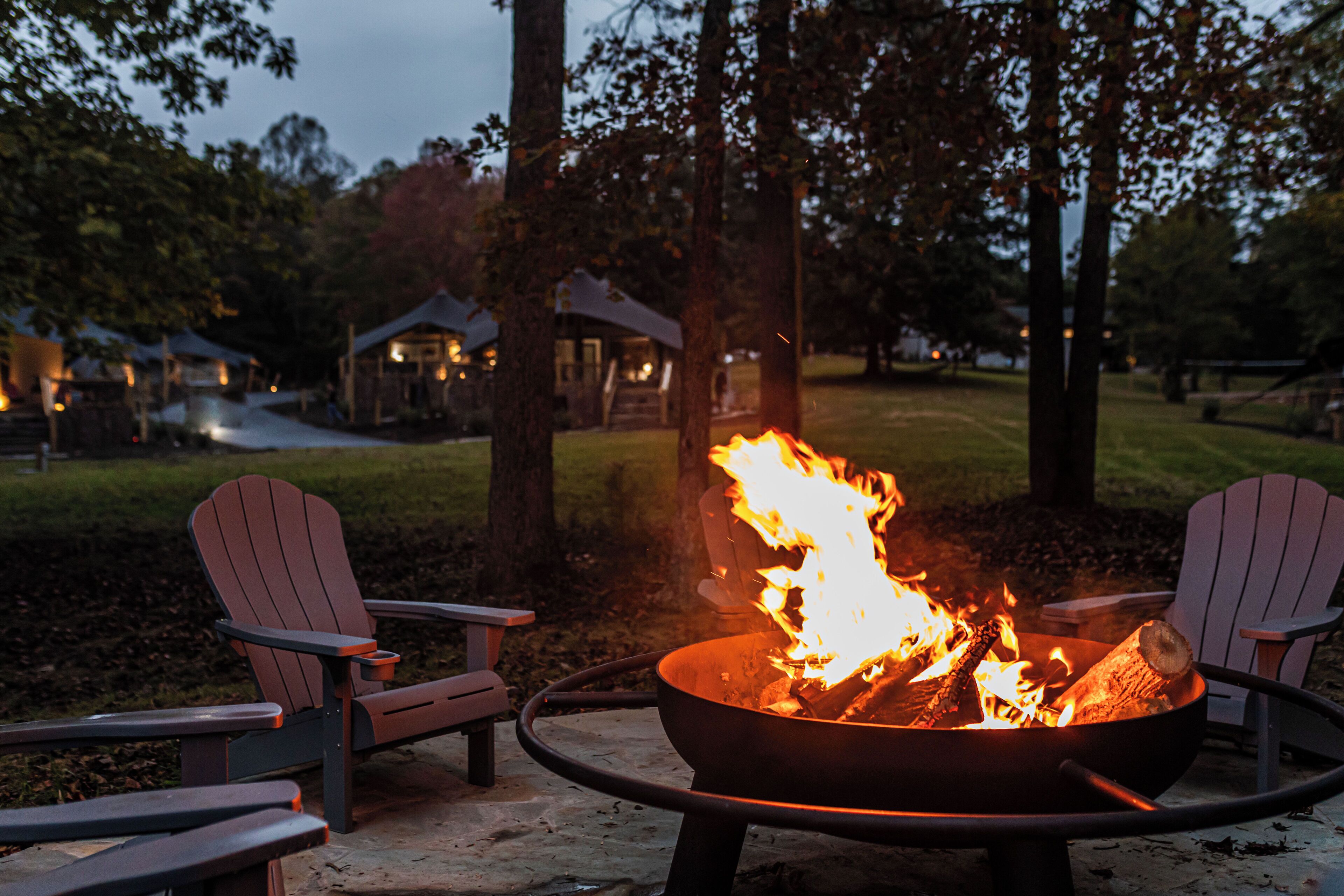 A fire pit with burning logs surrounded by Adirondack chairs in an outdoor setting.