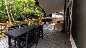 A covered outdoor deck with a table and chairs, surrounded by a wooden railing and a view of trees.