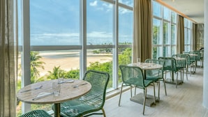 Un restaurante con amplios ventanales que ofrecen vista a una playa. Mesas y sillas dispuestas para comer.