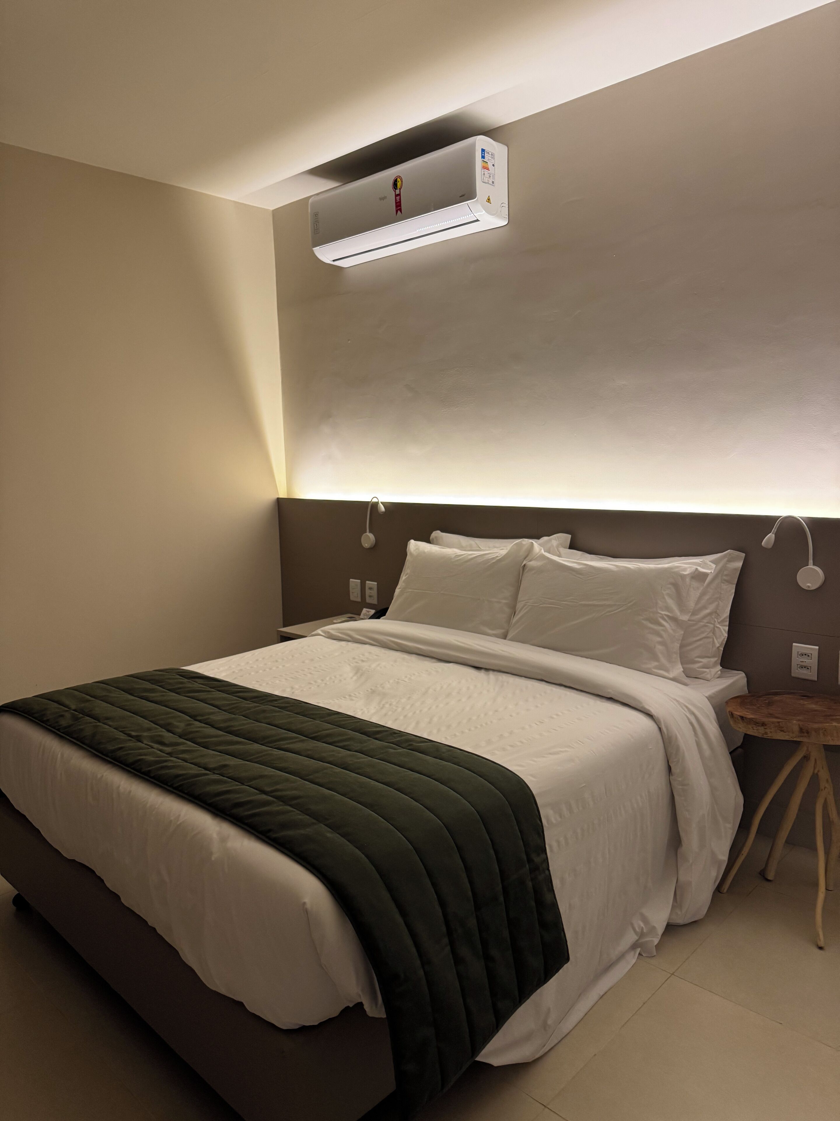 A hotel room with a bed, a bedside table, a nightstand, and a wall-mounted air conditioner.