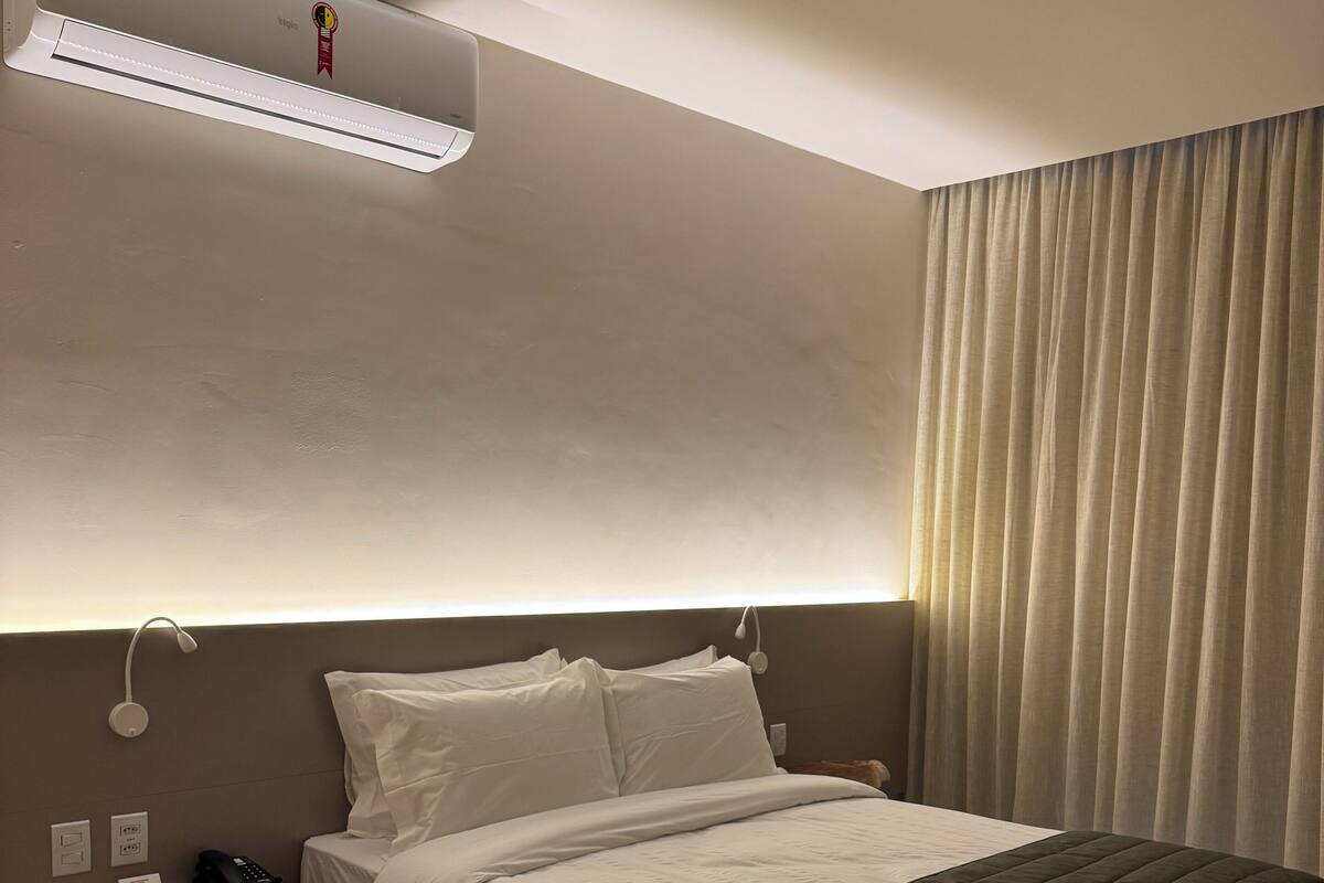 Una habitación de hotel moderna con una cama grande, una mesita de noche con teléfono y control remoto, y un equipo de aire acondicionado sobre la cama.