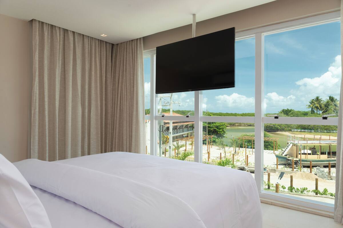 Habitación de hotel con cama, televisor montado en la pared y un amplio ventanal con vistas a un parque y un barco.