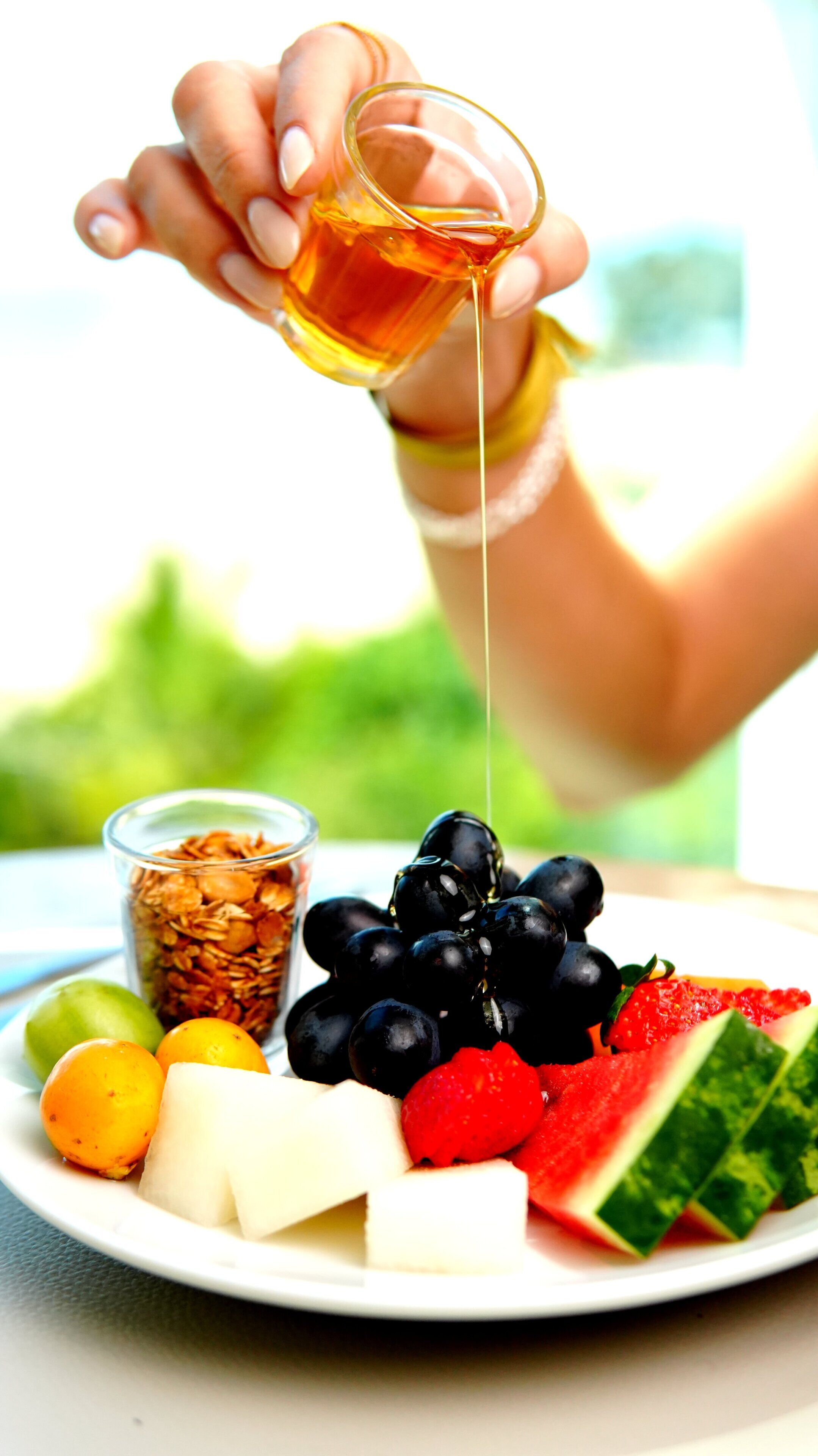 Una mano vertiendo miel sobre un plato con diversas frutas y queso.