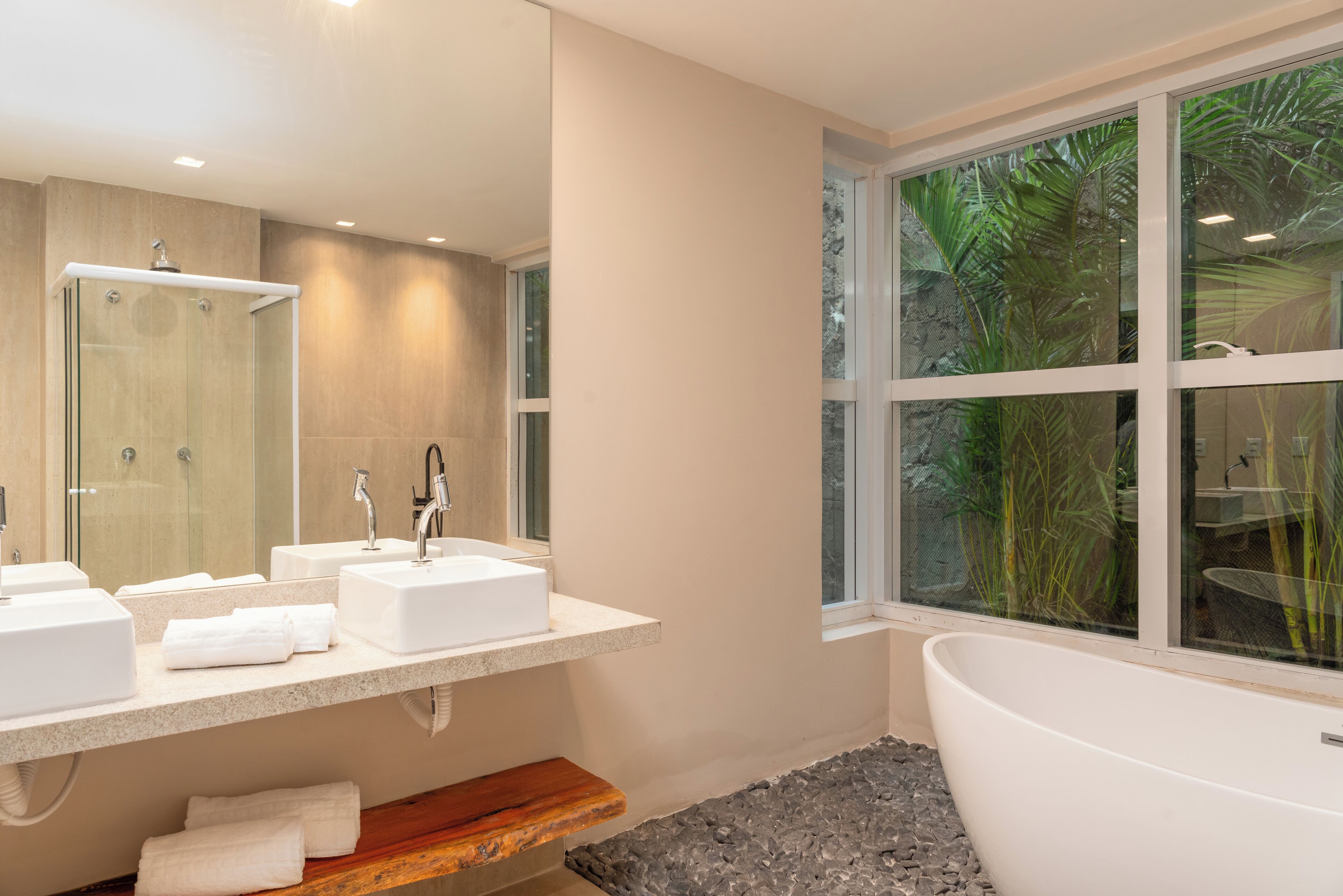 Ein modernes Badezimmer mit einer großen Badewanne, zwei Waschbecken, einer Duschkabine aus Glas und einem Fenster mit Blick auf üppige tropische Vegetation.