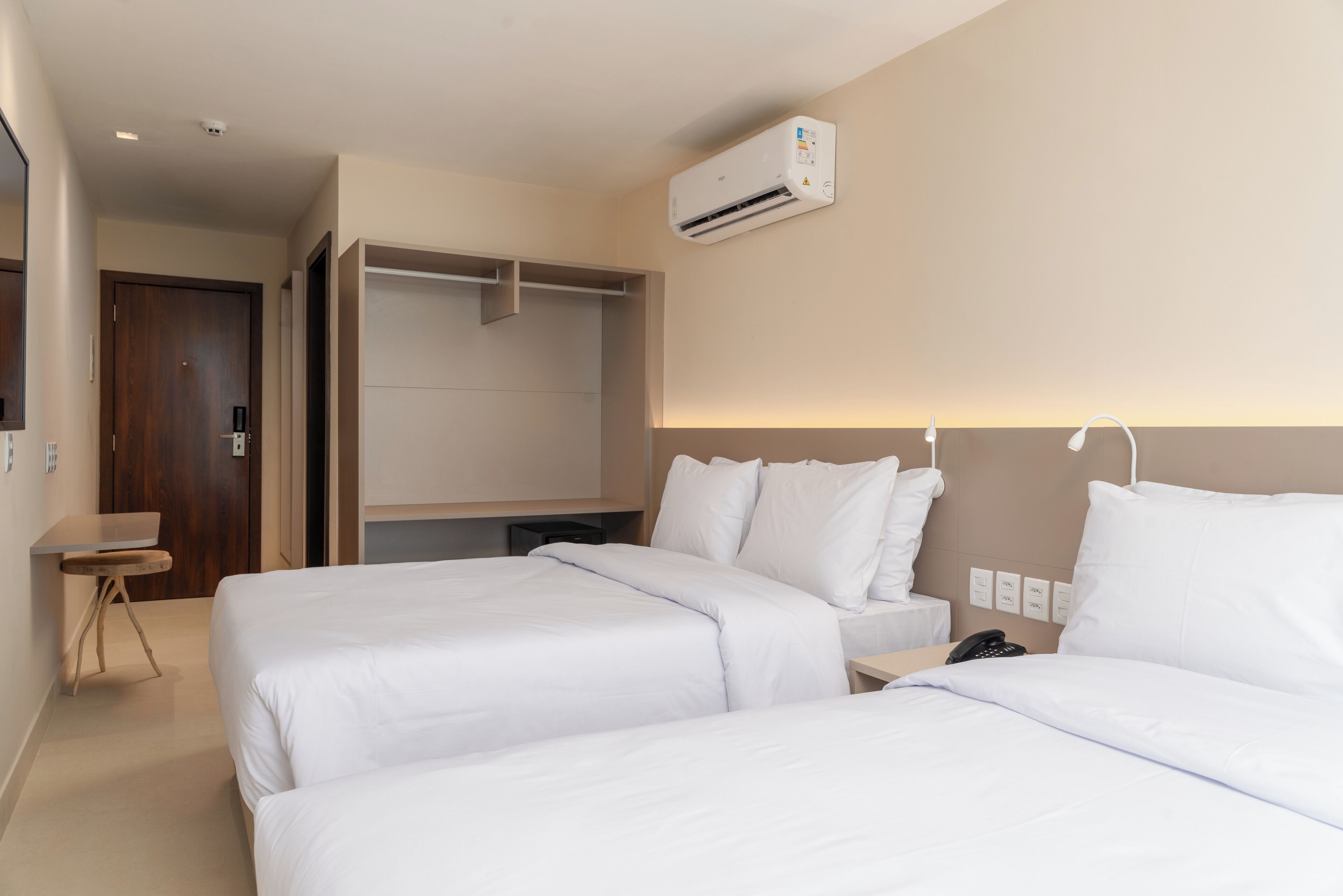 A hotel room with two beds, a desk, a wardrobe, and an air conditioning unit.