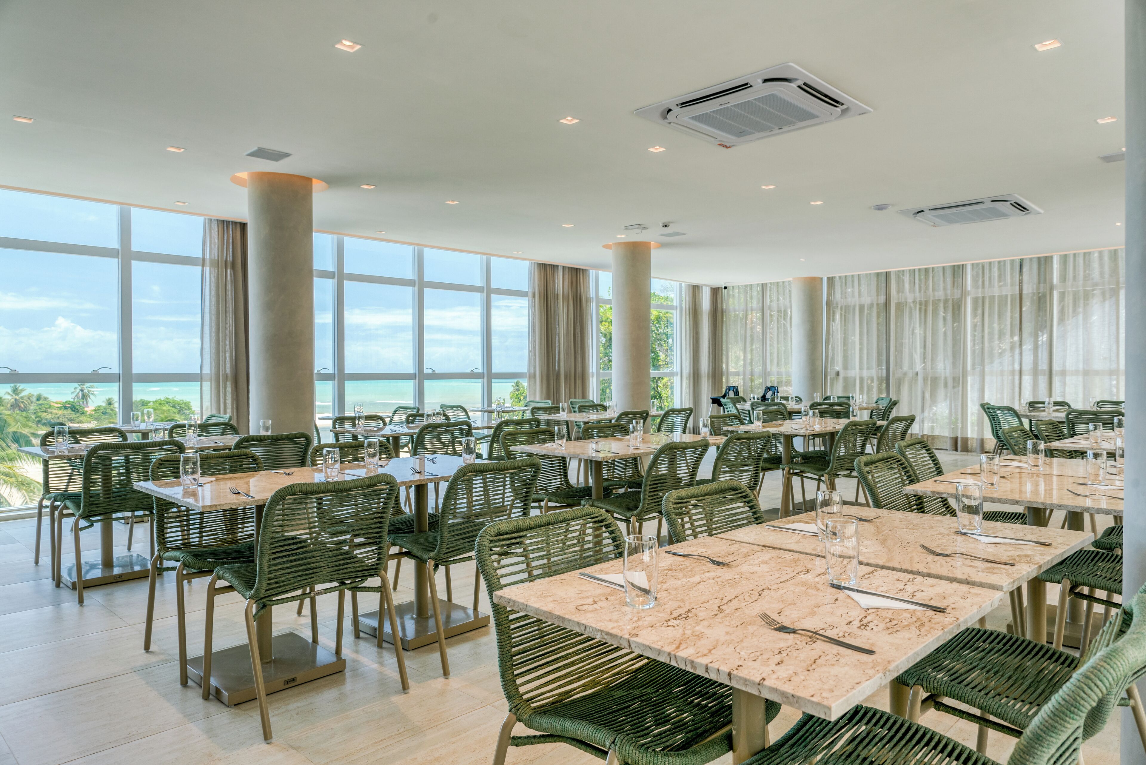 A spacious dining area with multiple tables and chairs, large windows offering a view of the ocean, and air conditioning units on the ceiling.