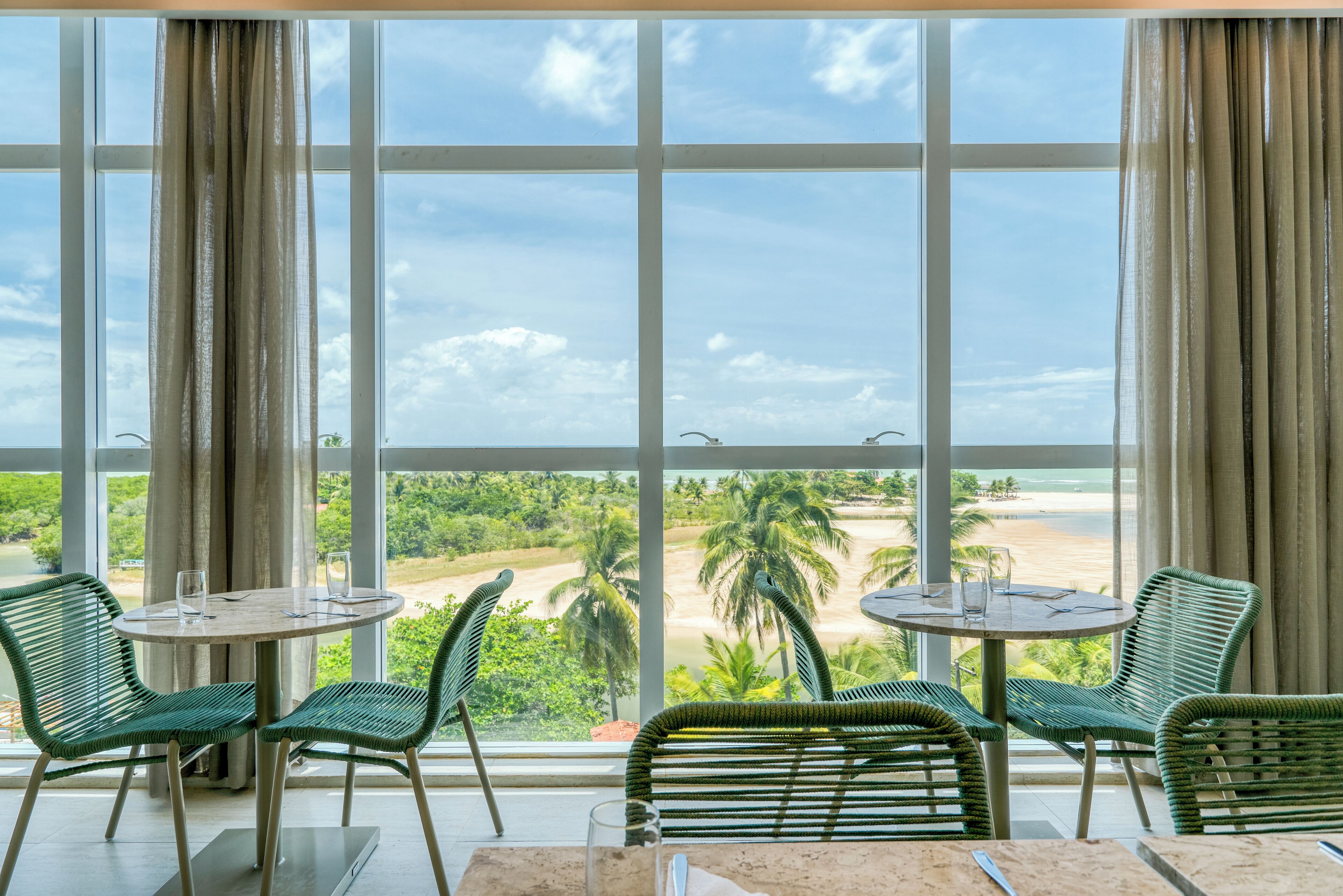 A room with large windows offering a view of a beach and palm trees. There are two round tables with chairs.