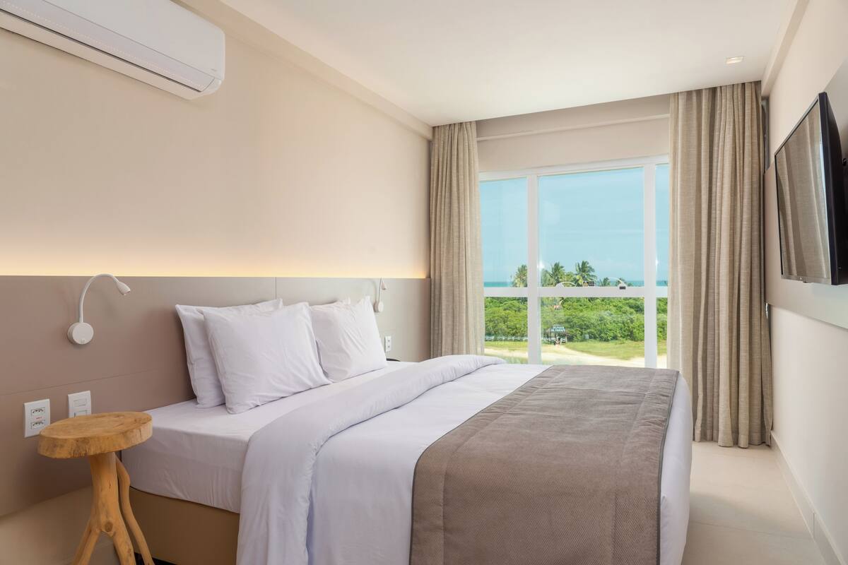 Una habitación de hotel moderna con una cama grande, una mesita de noche de madera, un televisor montado en la pared y un ventanal con vistas al océano.