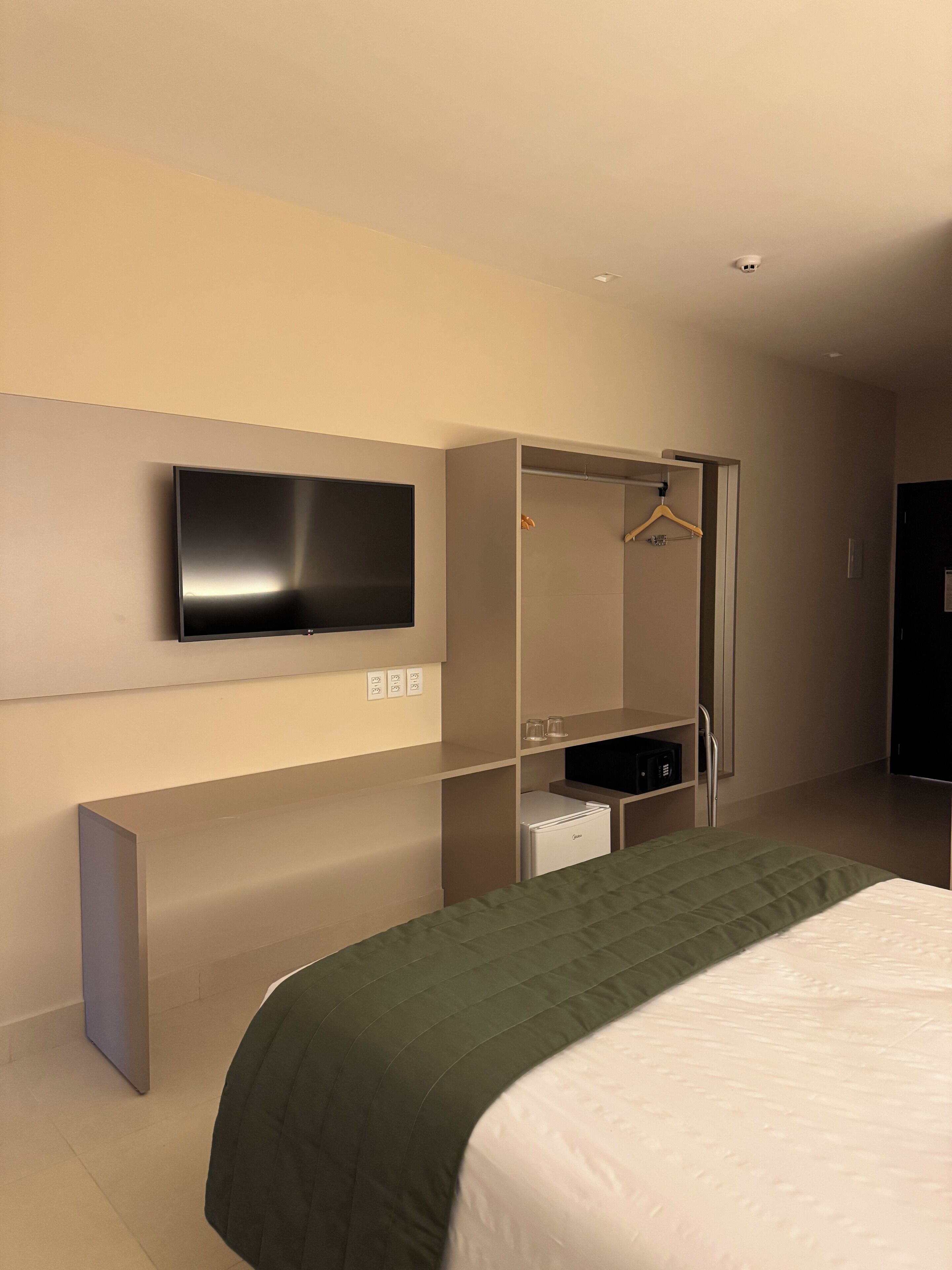 Una habitación de hotel moderna con una cama grande, un televisor de pantalla plana montado en la pared, un armario con espejo y un refrigerador pequeño.