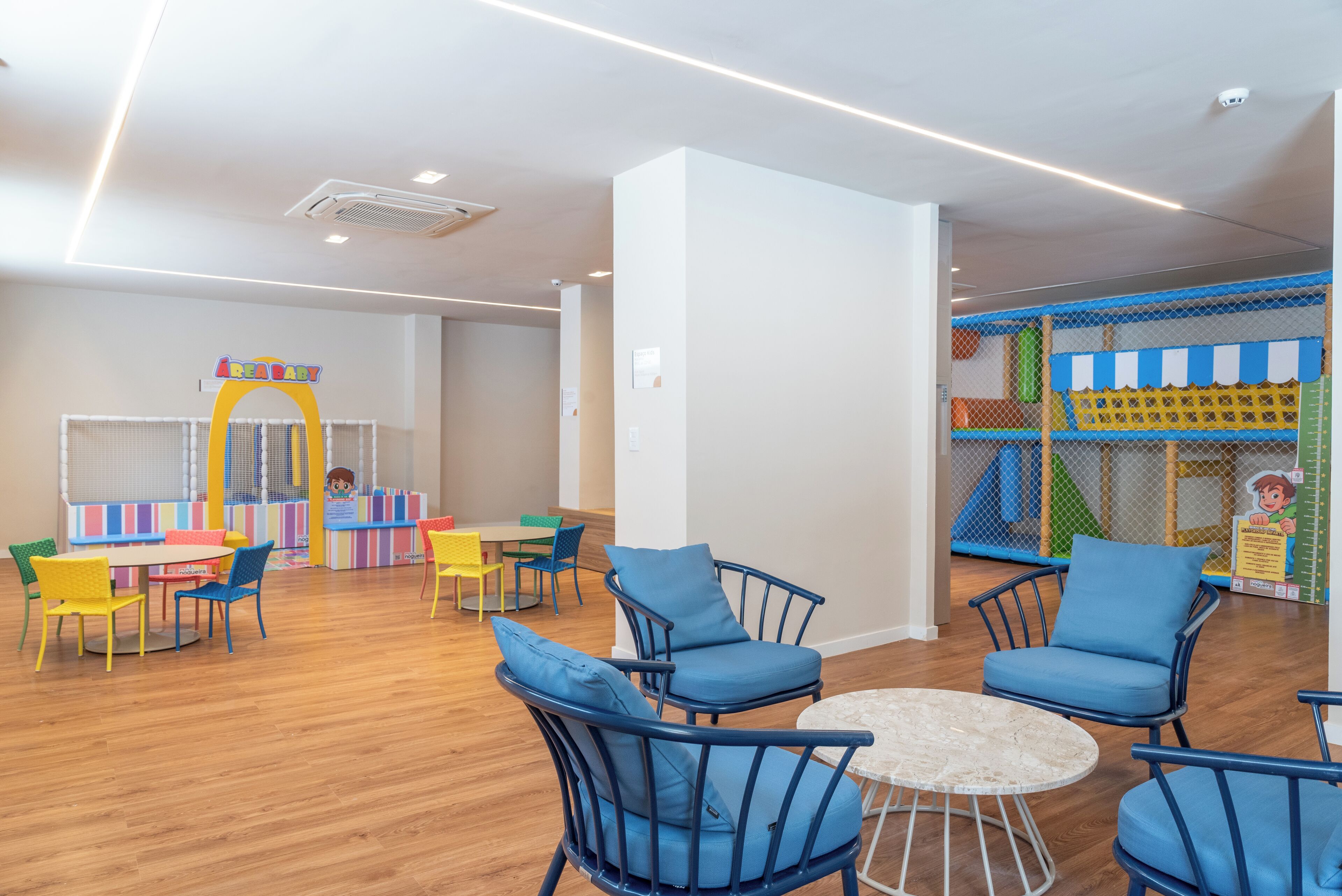 Ein Kinderspielbereich mit bunten Spielgeräten, einem Spielhäuschen und einer Sitzecke mit blauen Stühlen und einem kleinen runden Tisch.
