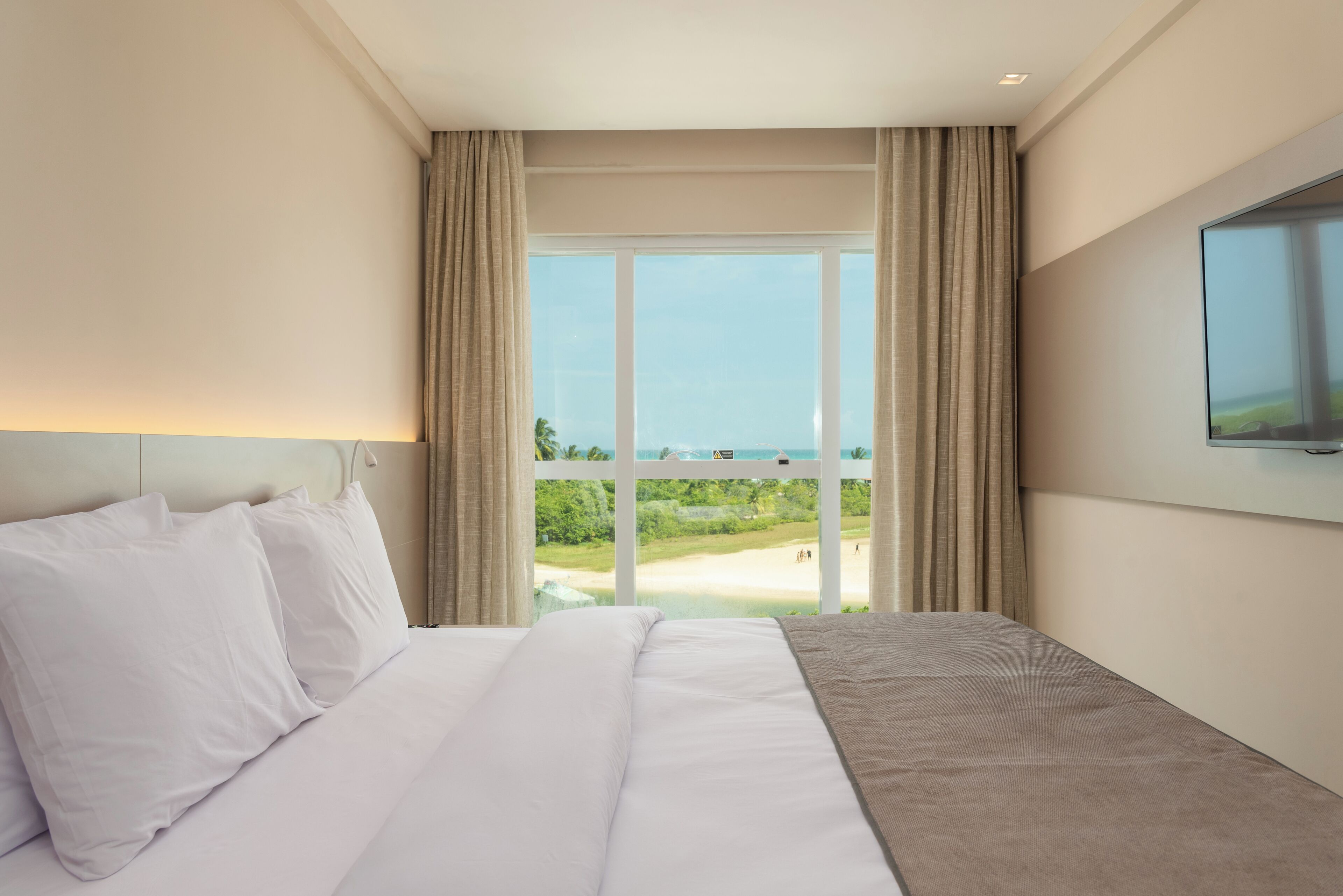 A hotel room with a large bed, a flat-screen TV mounted on the wall, and a view of a beach through a large window.