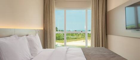 A hotel room with a large bed, a flat-screen TV mounted on the wall, and a view of a beach through a large window.