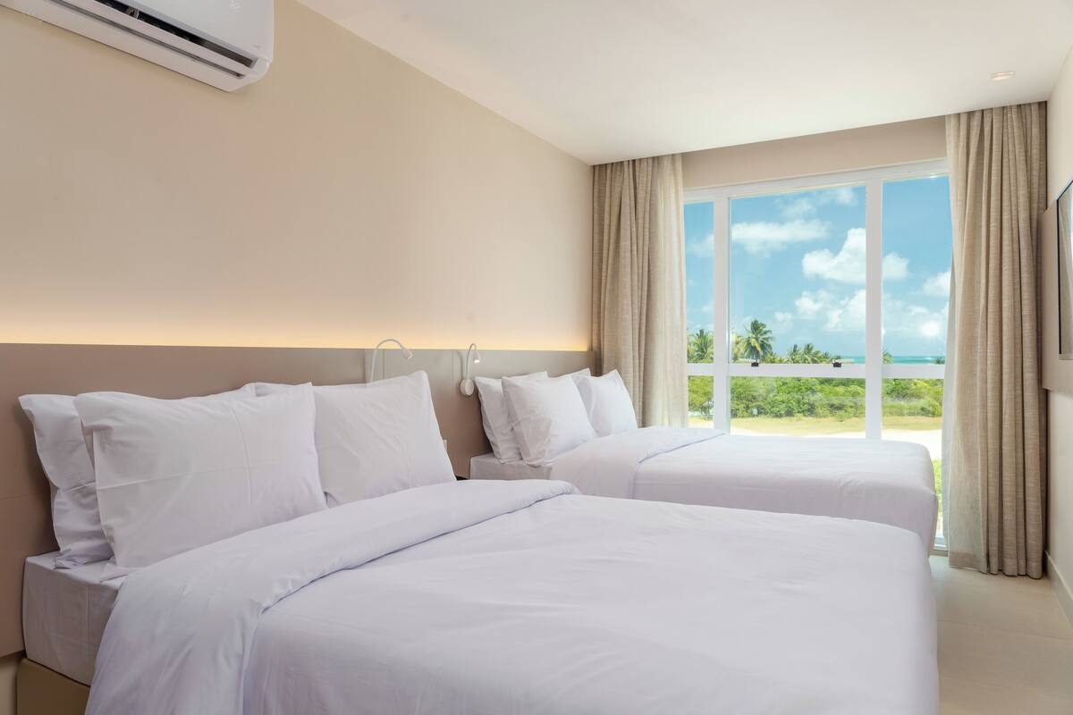 Habitación de hotel con dos camas, un ventanal con vista al océano y un equipo de aire acondicionado en la pared.