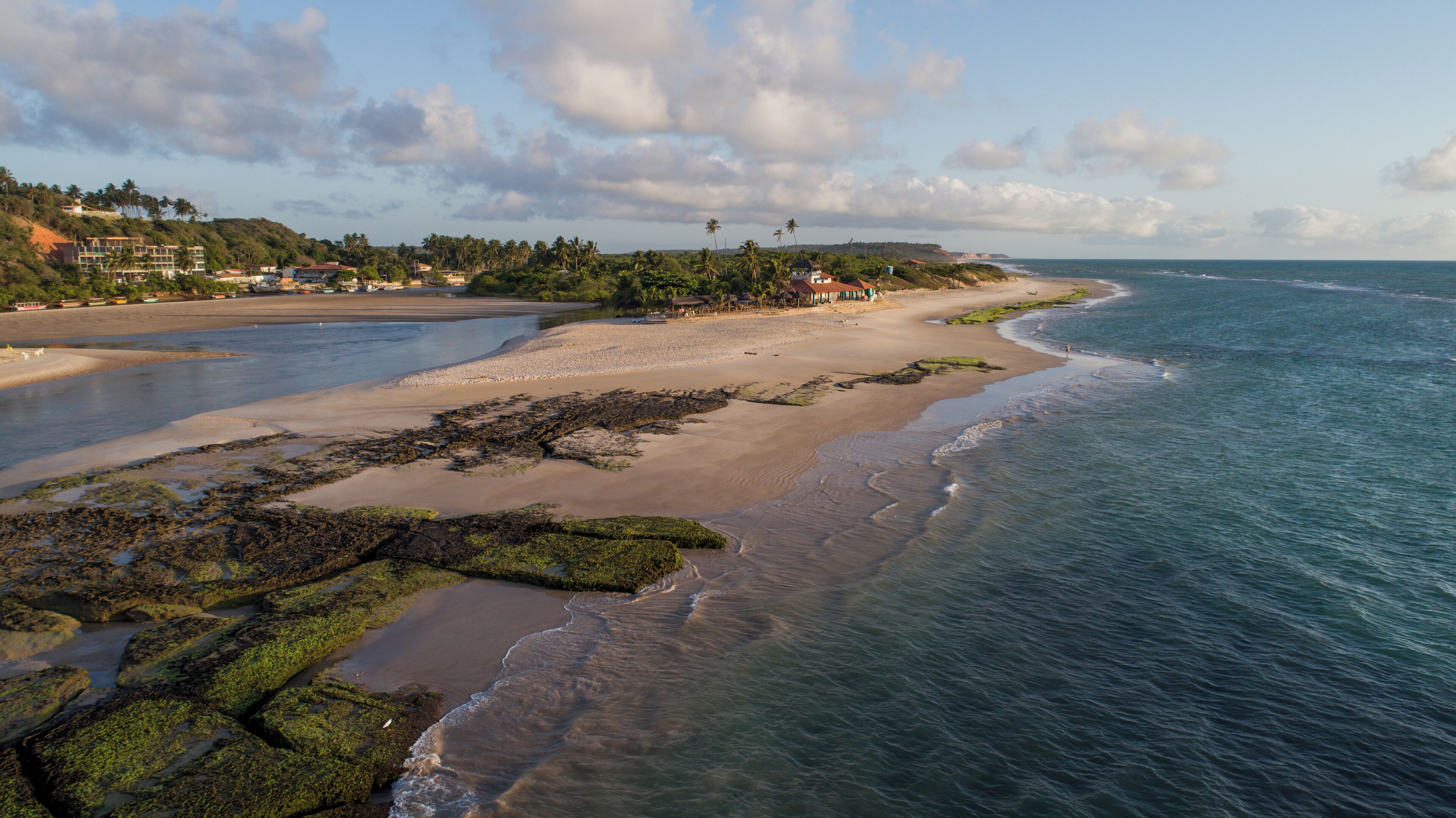 A coastal landscape with a beach, rocky shore, and a distant shoreline with buildings and vegetation.