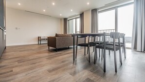 A spacious living area with a wooden floor, a sofa, a dining table with chairs, and large windows offering a view. - London East Village Apartment With City Views (London)