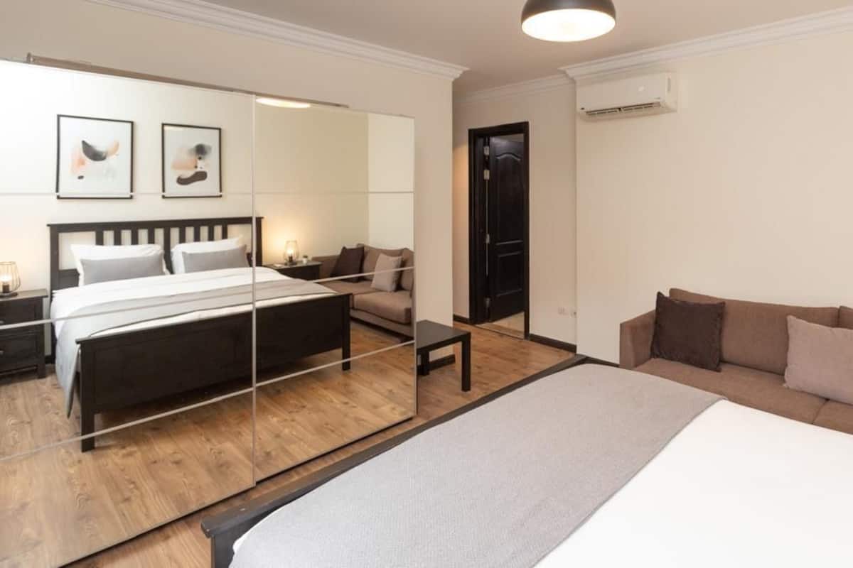 A modern bedroom with a large bed, a sofa, a bedside table, and a mirror.