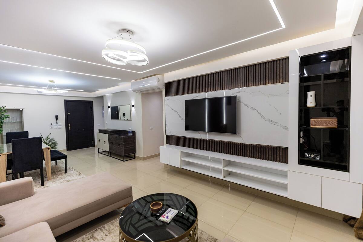 A modern living room with a flat-screen TV, a sofa, a coffee table, and a dining area with chairs and a table.